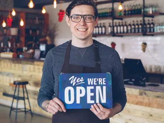 A cafe worker in an apron holding a sign that says Yes, we&rsquo;re open.