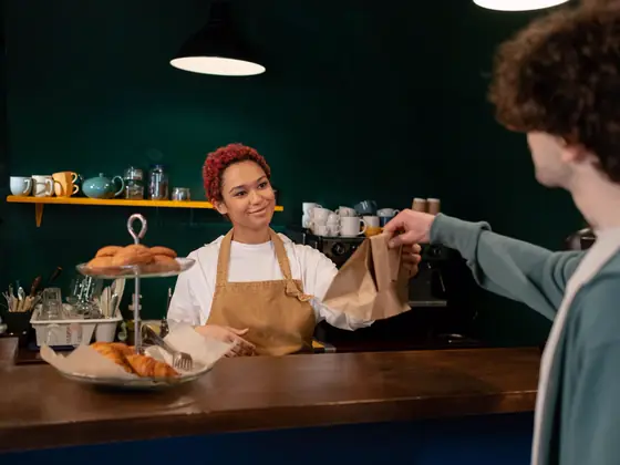 A cafe owner handing a customer a paper bag across the counter.