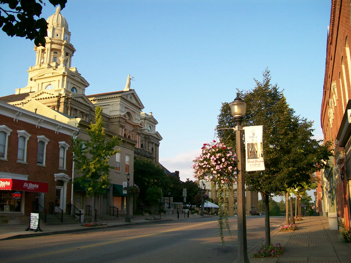 Main Street in downtown St. Clairsville at evening: brick storefronts, lamp posts with hanging flower baskets, and the courthouse tower down the street.