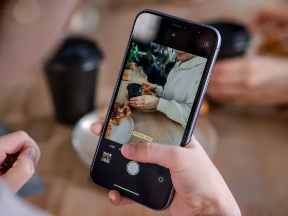 A hand holding a phone, filming a cafe table with coffee and a pastry.