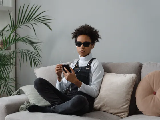 A young person in dark glasses on a sofa, listening to a phone through earphones.