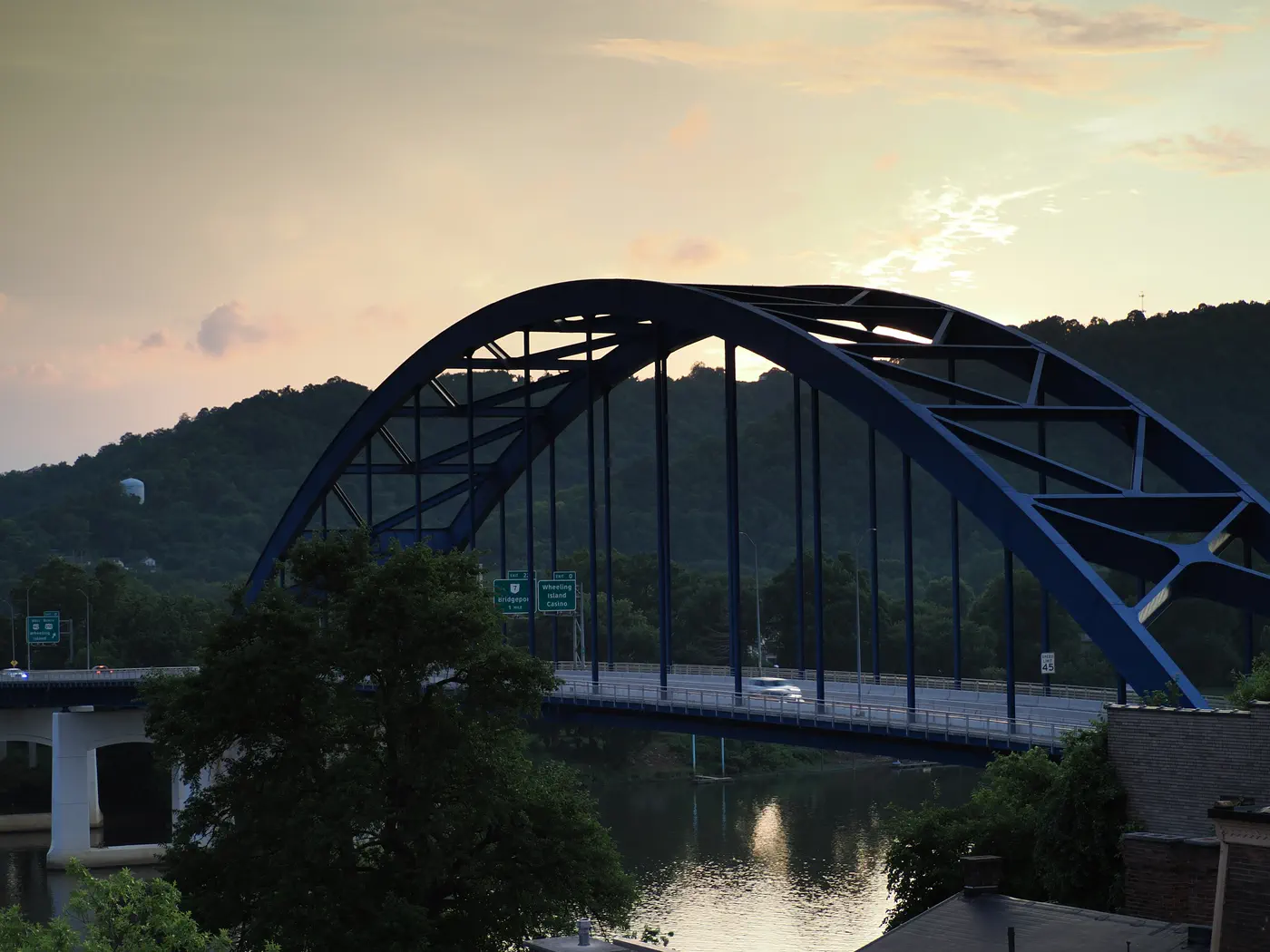 The royal blue arch of the Fort Henry Bridge over the Ohio River at dusk, the wooded hills behind.