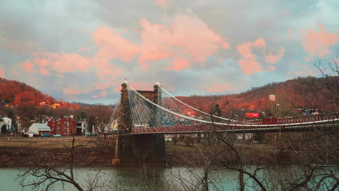 The Wheeling Suspension Bridge: its stone tower and cables over the Ohio River, pink clouds above the hills.