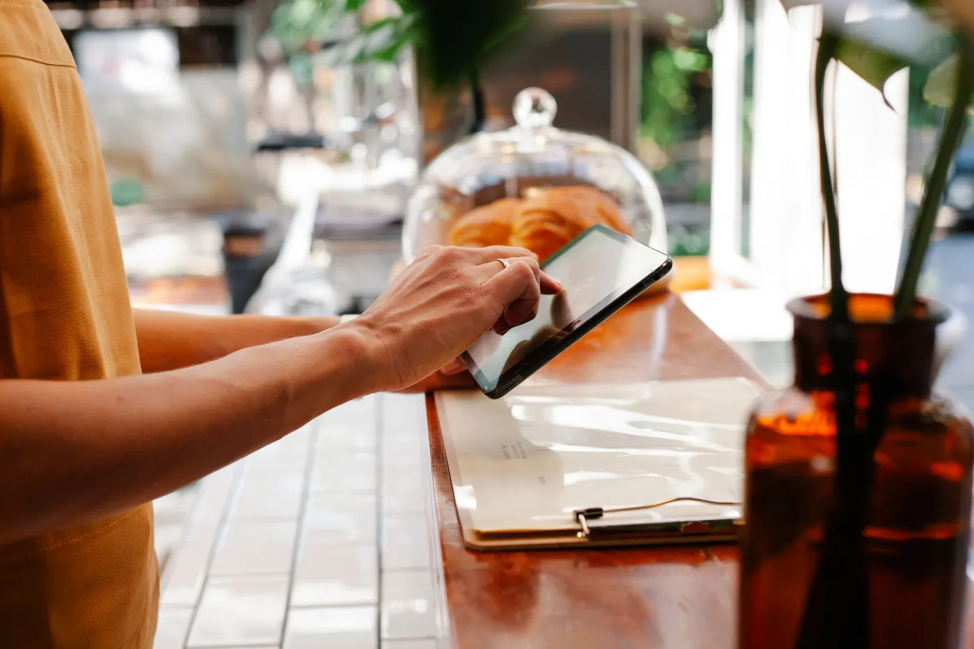 Hands tapping a tablet on a wooden cafe counter, beside a clipboard and a glass dome of croissants.