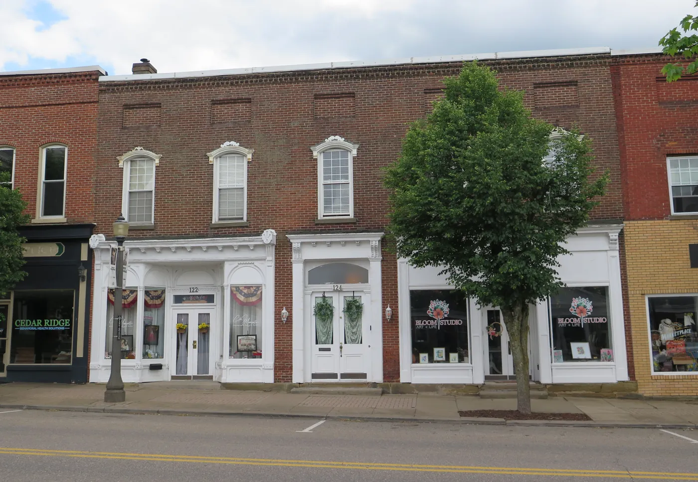 Brick storefronts on West Main Street in St. Clairsville, with white shopfronts, shop signs in the windows, a street lamp and a young tree.